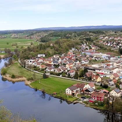 Panoramablick auf das Dorf Kallmünz mit rotgedeckten Häusern entlang eines Flusses, umgeben von grünen Wiesen und fernen Hügeln unter blauem Himmel.