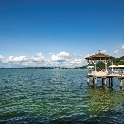 Wooden pier with white gazebo extending into Lake Constance in Bregenz. Clear blue sky with white clouds above the lake and forested hills.