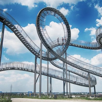 The Tiger and Turtle art installation in Duisburg featuring a large metallic roller coaster-like walkway with loops against a blue cloudy sky.