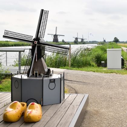 Kinderdijk Windmühlenmodell mit Holzschuhen Kleines Windmühlenmodell auf Holzplattform mit gelben Holzschuhen, Radfahrer auf Weg, historische Kinderdijk Windmühlen im Hintergrund.