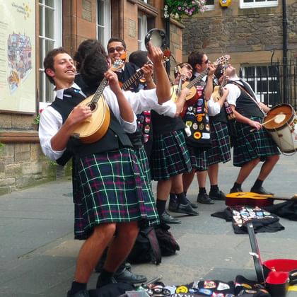 Group of Scottish street musicians performing in traditional green tartan kilts, playing mandolin, guitar and drums on a stone street.