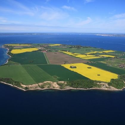 Luftaufnahme der Insel Ven, umgeben von blauem Meer. Die Insel zeigt Flickenteppich-Felder in Grün und Gelb, verstreute Gebäude und eine felsige Küste.