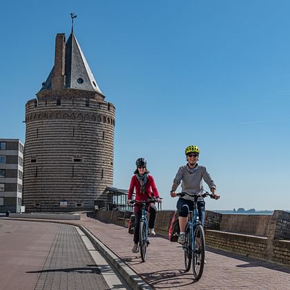 Two cyclists riding on the waterfront promenade in Vlissingen with a historic round tower and modern buildings under a clear blue sky.