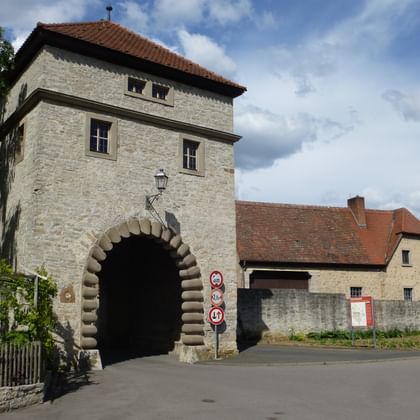 Mittelalterlicher Steinturm in Sommerach mit Torbogen, rotem Ziegeldach und traditionellen deutschen Häusern im Hintergrund unter bewölktem Himmel.