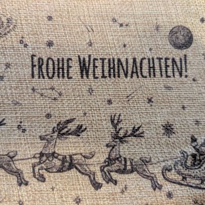Burlap fabric with 'Frohe Weihnachten' text and winter scene showing three reindeer pulling Santa's sleigh under a moon and snowflakes.