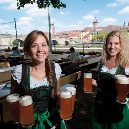 Zwei lächelnde Bedienungen in traditionellen Dirndl-Kleidern tragen Biergläser im Würzburger Biergarten mit historischer Brücke im Hintergrund.