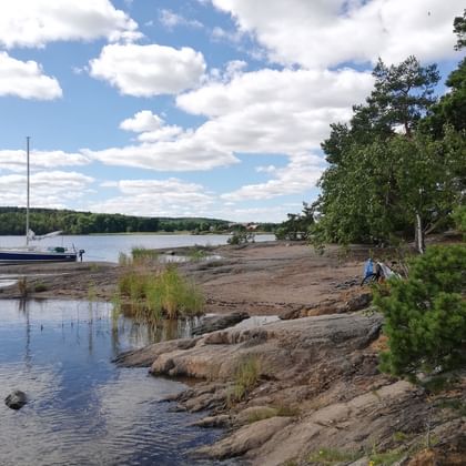 Rocky shoreline at Erstavik Nature Park with sailboat anchored in calm water. Pine trees line the granite coast under cloudy blue sky.