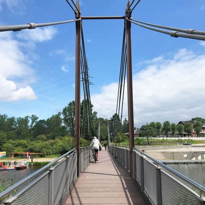 Cyclist crossing a suspension footbridge over the Elbe River in Geesthacht. The bridge has metal cables and wooden deck with green trees and harbor visible.