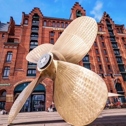 Large golden ship propeller sculpture in front of red brick warehouse buildings in Hamburg's historic Speicherstadt district under blue sky.
