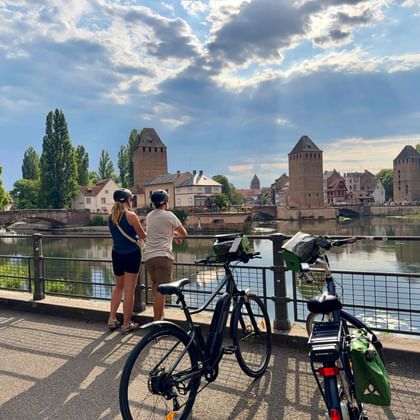 Zwei Radfahrer mit Rädern betrachten mittelalterliche Türme und Brücke über Fluss in Straßburg unter dramatischem Wolkenhimmel.