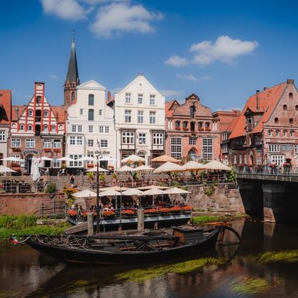 Historische Lüneburger Wasserfront mit bunten mittelalterlichen Gebäuden, Kirchturm, Steinbrücke und traditionellem Kanalboot im Vordergrund.