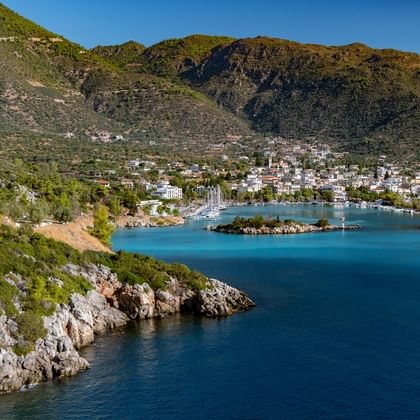 Scenic bay in Peloponnese with turquoise water, white buildings along the shore, rocky coastline, and green mountains under clear blue sky.