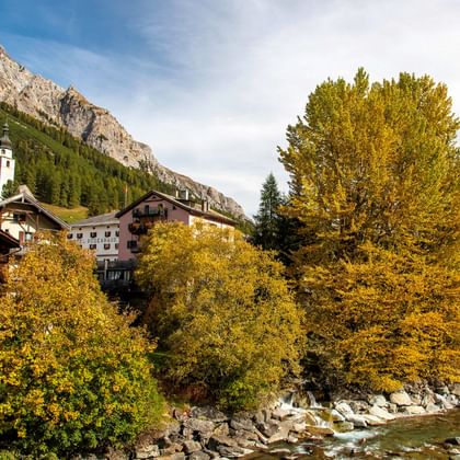 Alpine village of Splügen with traditional wooden chalets, white church tower, and golden autumn trees along a mountain stream.