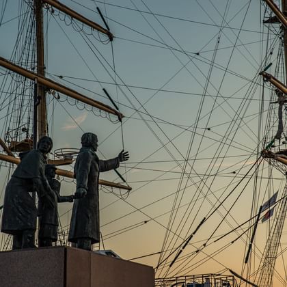 Bronze statue of two emigrants with luggage against historic sailing ship masts and rigging in Bremerhaven at sunset.