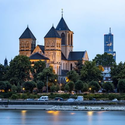 St. Kunibert Kirche mit Zwillingstürmen beleuchtet in der Dämmerung am Rhein in Köln, mit modernen Gebäuden im Hintergrund.