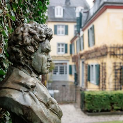 Bronzeskulptur von Beethoven mit lockigem Haar im Profil, umgeben von Efeu, mit gelben historischen Gebäuden im Hintergrund in Bonn.