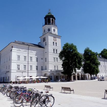 White baroque Salzburg Museum building with prominent clock tower on a sunny square. Bicycles are parked in front, benches line the plaza.