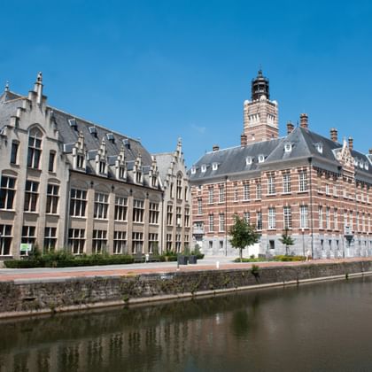 Row of historic Flemish buildings with ornate facades and towers reflected in calm river water under blue sky in Dendermonde, Belgium.