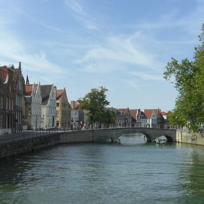 Historischer Kanal mit Brücke in Brügge Steinbrücke über einen Kanal in Brügge mit mittelalterlichen Gebäuden an beiden Seiten. Grüne Bäume rahmen die Szene unter teilweise bewölktem Himmel.