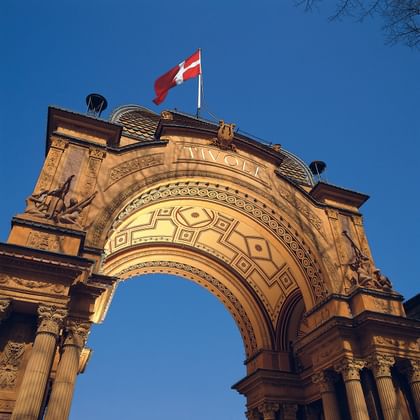 Ornate entrance arch of Tivoli Gardens with decorative patterns and the word 'TIVOLI' on top. A Danish flag flies above against a clear blue sky.