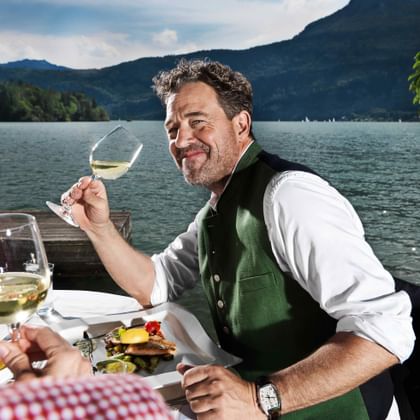 Man in traditional vest enjoying white wine at lakeside table with mountains in background at Zeller See, Salzburger Land, Austria.