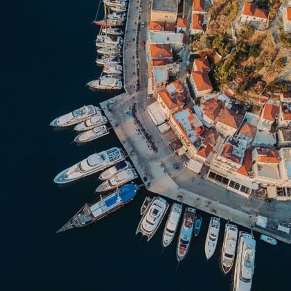 Luftaufnahme eines mediterranen Hafens auf dem Peloponnes mit Booten entlang der geschwungenen Uferpromenade, traditionellen rotgedeckten Gebäuden.