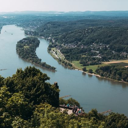 Luftaufnahme des Rheins, der sich durch grüne Landschaft bei Bonn schlängelt, mit bewaldeten Hügeln, Feldern und Besuchern am Aussichtspunkt.