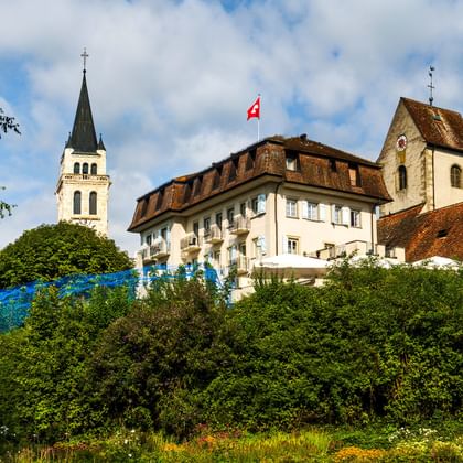 View of Romanshorn City Garden showing historic buildings with red tile roofs, a church spire, and Swiss flag, surrounded by lush green trees.