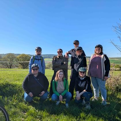 Group of nine cyclists posing in two rows on a grassy hillside with green fields and blue sky in the background.