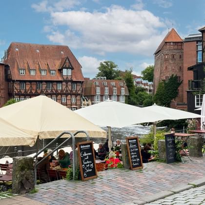 Waterfront café with white umbrellas at Lüneburg's Old Harbor. Historic half-timbered houses and brick tower visible behind cobblestone plaza.