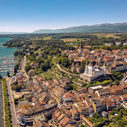 Aerial view of Nyon, a historic town on Lake Geneva with terracotta roofs, a castle, marina, and mountains in the background.