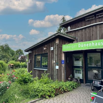 Wooden Boberger Dünenhaus building with green sign, surrounded by flowering garden. A bicycle trailer stands at the entrance under blue sky.