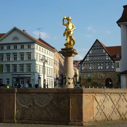 St. Georg Statue auf Eisenacher Marktplatz Goldene St. Georg Statue auf Steinsäule auf dem Eisenacher Marktplatz, umgeben von historischen Gebäuden mit Fachwerkhäusern und Kirche.