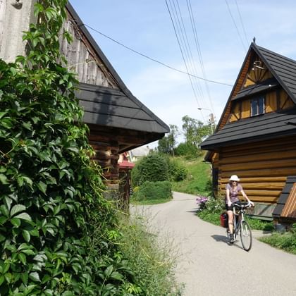 Cyclist riding through a Polish village with traditional wooden houses. Left side shows an old barn, right side a log house with steep roof.