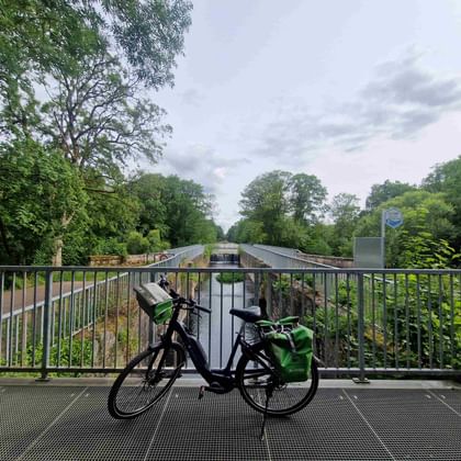 Fahrrad mit grünen Taschen auf einem Metallbrückengeländer. Bäume und eine Kanalschleuse im Hintergrund unter bewölktem Himmel.