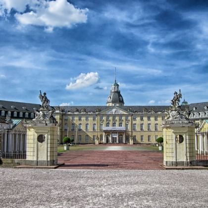 Karlsruhe Palace with yellow baroque facade and central tower, viewed through ornate entrance gates with sculptures under a dramatic blue sky.