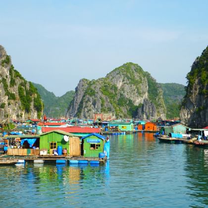 Bunte schwimmende Häuser und Boote auf ruhigem Wasser, umgeben von hohen Kalkstein-Karstformationen in Vietnam. Grüne und blaue Häuser.