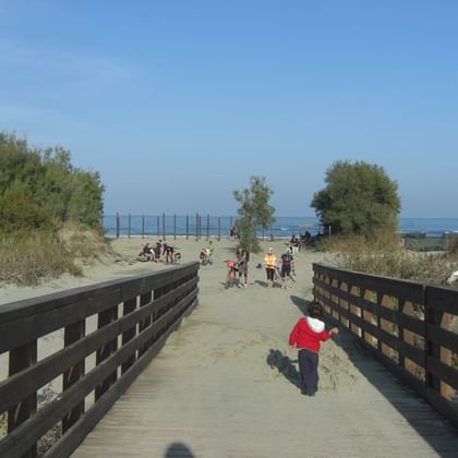 Wooden boardwalk leading to Boccasette Beach with visitors walking on sandy path. Child in red shirt walks on boardwalk toward beach area.