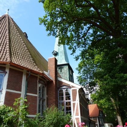 Red brick church with copper-green spire in Bergedorf, framed by large trees and pink flowers, under blue sky.