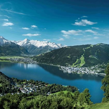 Aerial view of Zell am See-Kaprun with blue lake surrounded by green mountains and snow-capped Alpine peaks under a bright blue sky.