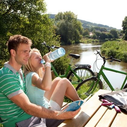 Cyclists resting by the Werra River Couple sitting on wooden bench by Werra River, woman drinking from water bottle. Green bicycle and cycling bags nearby, lush green landscape.