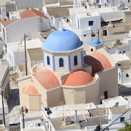 Luftaufnahme einer griechisch-orthodoxen Kirche mit blauer Kuppel und rot-orangen kleineren Kuppeln, umgeben von weißen kubischen Häusern auf Seriphos.