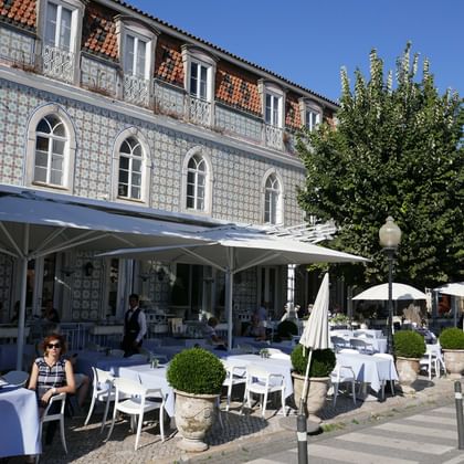 Outdoor cafe terrace in Sintra with white tables and chairs under umbrellas. Historic Portuguese building with azulejo tiles and arched windows.