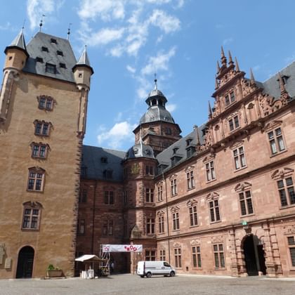 Innenhof von Schloss Aschaffenburg mit roten Sandstein-Renaissance-Gebäuden, runden Türmen, verzierten Fassaden und zentraler Kuppel unter blauem Himmel.