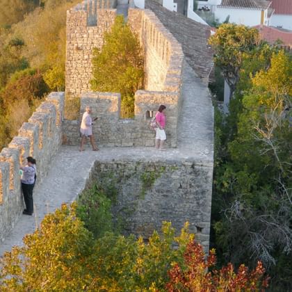 Aerial view of Castelo de Óbidos medieval stone walls with three visitors walking along the fortifications, surrounded by autumn trees.