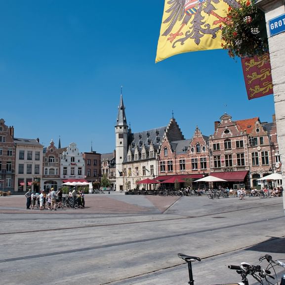 Historischer Marktplatz in Dendermonde mit bunten flämischen Gebäuden, gotischem Rathaus mit Uhrenturm, Straßencafés und Fahrrädern unter blauem Himmel.
