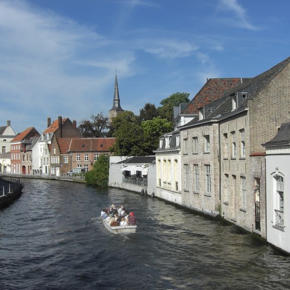 Gracht in Brügge mit Touristenboot voller Passagiere zwischen historischen Gebäuden. Mittelalterliche Architektur säumt beide Seiten.