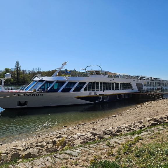 White river cruise ship named Manon moored near a rocky shoreline under clear blue sky. The multi-deck vessel has large windows and is on calm water.