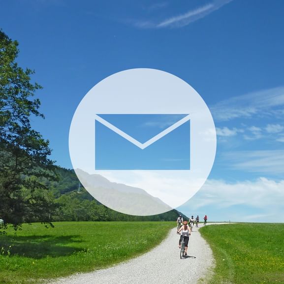Großes weißes Briefumschlag-Symbol über malerischer Landschaft mit Radfahrern auf Schotterweg durch grüne Felder unter blauem Himmel.