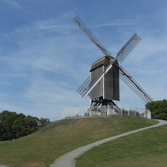 Sint Janshuismolen, eine traditionelle Holzwindmühle mit vier Flügeln, steht auf einem Grashügel umgeben von Bäumen unter blauem Himmel.